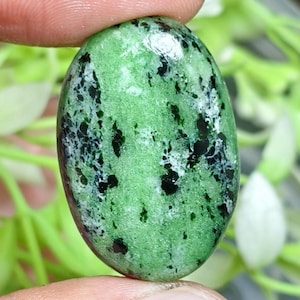 Op de afbeelding: Een ovaalvormige cabochon edelsteen met een levendige groene basiskleur en opvallende zwarte en witte gespikkelde insluitingen over het gepolijste oppervlak. De steen heeft een gladde, afgeronde bovenkant en een glanzende afwerking.