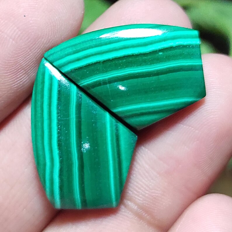May include: Two polished malachite gemstones, cut into geometric shapes, are displayed. The stones are a vibrant green with darker green bands, creating a striped pattern. The gemstones are held in a hand.