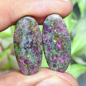 Peut inclure: Deux cabochons de forme ovale présentant un motif tacheté d'inclusions cristallines magenta foncé et violettes incrustées dans une matrice vert clair, avec une surface polie lisse et des textures minérales naturelles et veines brunes.
