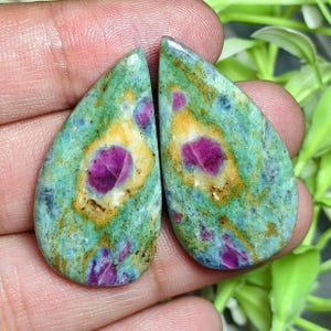 Op de afbeelding: Twee peervormige cabochons met een mix van groene, blauwe en gele mineraalpatronen met duidelijke, ingebedde magentakleurige robijninsluitingen. De stenen hebben een glad, gepolijst oppervlak en een druppelvormig silhouet.