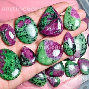 May include: A collection of polished ruby zoisite gemstones in various shapes including ovals, teardrops, and irregular forms. Each stone features a distinct mottled pattern of vibrant magenta-pink ruby inclusions set against a green zoisite base.