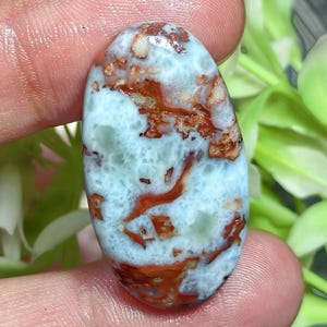 Puede incluir: Una piedra preciosa cabujón de forma ovalada con una superficie pulida que presenta un patrón jaspeado en tonos azul cielo y blanco, intercalado con vetas irregulares y parches de inclusiones minerales de color marrón rojizo.