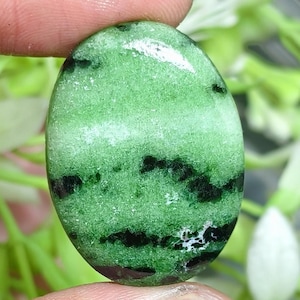 Puede incluir: Una piedra preciosa en cabujón de forma ovalada que presenta un color base verde claro vibrante con prominentes inclusiones minerales irregulares de color verde oscuro a negro distribuidas por la superficie, pulida con un acabado liso, brillante y reflectante.