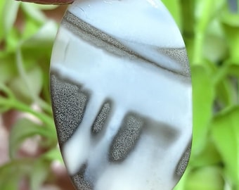 May include: An oval-shaped polished gemstone featuring a smooth white surface with distinct, textured gray inclusions that form horizontal bands and vertical, drip-like patterns across the stone's face, creating a high-contrast natural appearance.
