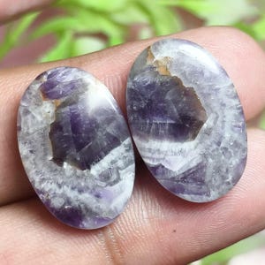 Puede incluir: Dos piedras preciosas de amatista de forma ovalada con una superficie pulida que presenta una mezcla de inclusiones de color púrpura intenso, blanco translúcido y marrón. Las piedras muestran una estructura cristalina natural con fracturas.
