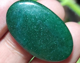 May include: A polished, oval-shaped gemstone with a deep emerald green color. The stone has a smooth, reflective surface with subtle variations in tone and texture, suggesting natural inclusions. The gemstone is held in a hand.
