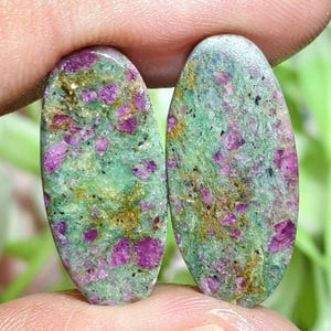 Pode incluir: Dois cabochões de formato oval com uma base manchada em verde e verde claro, com inclusões de cristais magenta vibrantes e veios minerais castanho-dourados espalhados pelas superfícies de pedra polida.