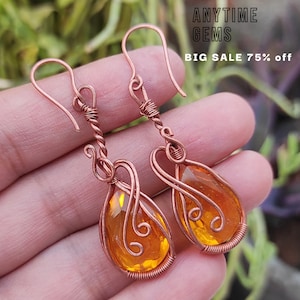 May include: A pair of copper wire earrings with orange teardrop-shaped glass beads. The earrings are handmade and feature a delicate, intricate design.