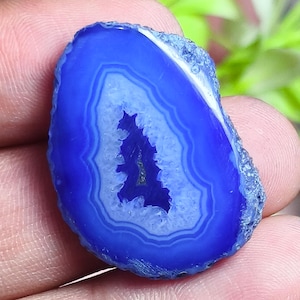 May include: A polished, vibrant blue agate slice featuring concentric bands of varying blue shades and a central, jagged geode cavity lined with small, sparkling crystals. The outer edge of the stone is rough, textured, and dark blue in color.