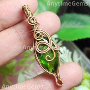 May include: A handcrafted pendant with a faceted, emerald-green gemstone set in intricate, antique-bronze wire. The wirework forms decorative swirls and wraps around the gemstone, creating a unique design. This pendant is a piece of jewellery.