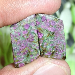 Peut inclure: Deux pierres précieuses polies de forme trapézoïdale présentant un mélange vibrant de cristaux de rubis rose magenta incrustés dans une matrice de zoïsite vert clair. Les pierres présentent une texture naturelle tachetée avec des inclusions minérales brun foncé.