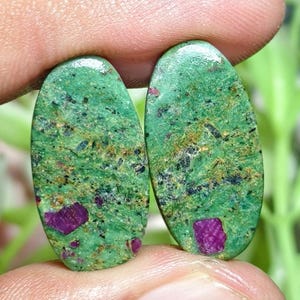 Peut inclure: Deux cabochons de forme ovale présentant une base vert vif avec des inclusions texturées noires et dorées. Chaque pierre polie affiche une inclusion cristalline distincte de couleur magenta profond incrustée dans la matière verte.