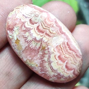 May include: An oval-shaped gemstone with a swirling pattern of pink, cream, and light brown hues. The polished surface displays a unique, layered design, with a mix of wavy and feathery textures. The stone is held in a hand, showcasing its natural beauty.