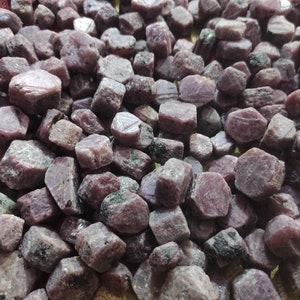 May include: A close-up view of a pile of rough, purple-hued ruby gemstones. The stones are various sizes and shapes, with some showing a hexagonal pattern.