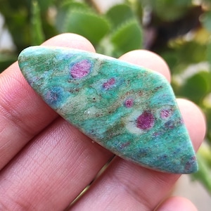May include: A polished gemstone with a green base color, speckled with vibrant magenta and blue hues. The stone has a smooth, elongated shape, and the magenta spots are scattered throughout the surface.