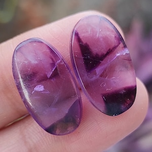 May include: Two oval-shaped, polished gemstones in a deep purple hue and translucent quality. The stones have a mix of dark and light purple shades, with some areas appearing almost black. The gemstones are likely amethyst, often used in jewellery.