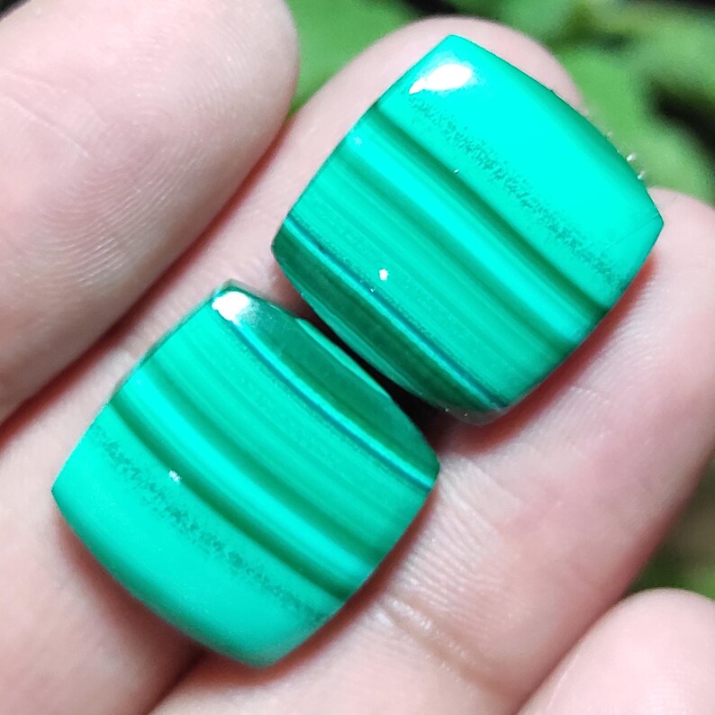 May include: Two square-shaped malachite gemstones with a polished finish. The stones display a vibrant green color with darker green bands and patterns. The gemstones are held in a hand.