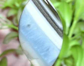 May include: A marquise-shaped cabochon gemstone featuring a smooth, polished surface with distinct horizontal bands of pale blue, opaque white, and a dark, shimmering metallic gray stripe running diagonally across the upper section.