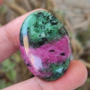 May include: An oval gemstone with a polished surface. The stone displays a vibrant mix of colours, including deep red, green, and black. The red and green hues are the most prominent, creating a striking contrast. The gemstone is likely a ruby in zoisite.