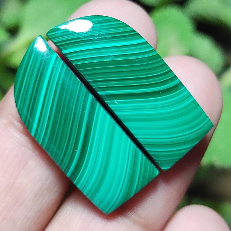 May include: Two polished malachite gemstone cabochons. The stones are a vibrant green with distinctive banded patterns. The cabochons are cut into a heart shape, with a flat back and a rounded top. The malachite has a glossy finish.
