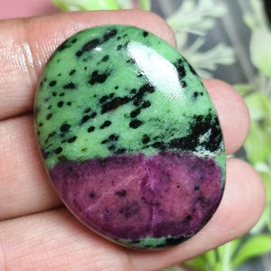 Puede incluir: Una piedra preciosa pulida de forma ovalada con una clara división de colores. La parte superior es de color verde claro con motas negras dispersas, mientras que la sección inferior es de un magenta púrpura intenso con inclusiones oscuras.