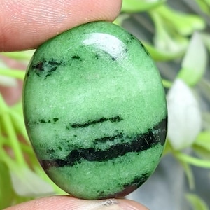 Puede incluir: Una piedra preciosa cabujón de forma ovalada con una superficie lisa y pulida de color base verde claro vibrante, acentuada por distintas vetas horizontales y parches de inclusiones minerales de color negro intenso.