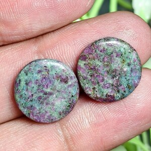 Peut inclure: Deux cabochons circulaires et plats présentant un motif tacheté d'inclusions minérales magenta profond et vert vif. Les pierres ont une surface polie et lisse avec une finition brillante, montrant une texture naturelle mouchetée.
