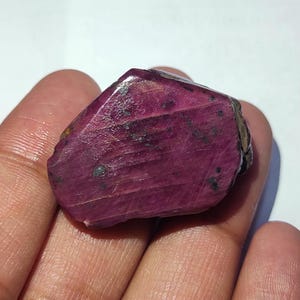 May include: A large, deep red ruby crystal with a rough, faceted surface. The crystal has a dark red color with black inclusions.