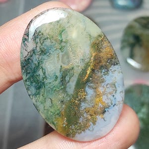 Puede incluir: Una piedra preciosa de ágata musgosa ovalada y pulida. La piedra presenta un patrón natural de inclusiones verdes, doradas y blancas, que se asemeja a un paisaje. El ágata se sostiene en una mano, mostrando su tamaño y detalles.
