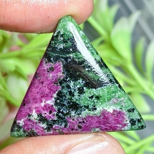 Puede incluir: Una piedra preciosa pulida de forma triangular que presenta una mezcla vibrante de inclusiones minerales en magenta intenso, verde bosque y negro. La superficie es lisa y reflectante, mostrando un patrón natural moteado con zonas de color distintas.
