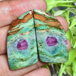 Puede incluir: Dos cabujones semicirculares a juego que presentan una vibrante matriz verde y marrón con una prominente inclusión de piedra preciosa de color magenta en el centro. Las superficies pulidas muestran patrones de remolinos en tonos verde azulado, menta y marrón tierra.