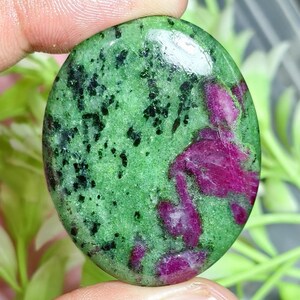Puede incluir: Una piedra preciosa pulida de forma ovalada que presenta una base de color verde vibrante con inclusiones prominentes de color magenta rosado intenso y motas minerales negras dispersas por toda la superficie lisa y brillante.