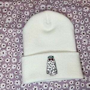 Pode incluir: Bon&eacute; branco com um fantasma bordado em branco que usa uma coroa de folhas verdes e bagas vermelhas. O fantasma tem olhos pretos e uma boca.