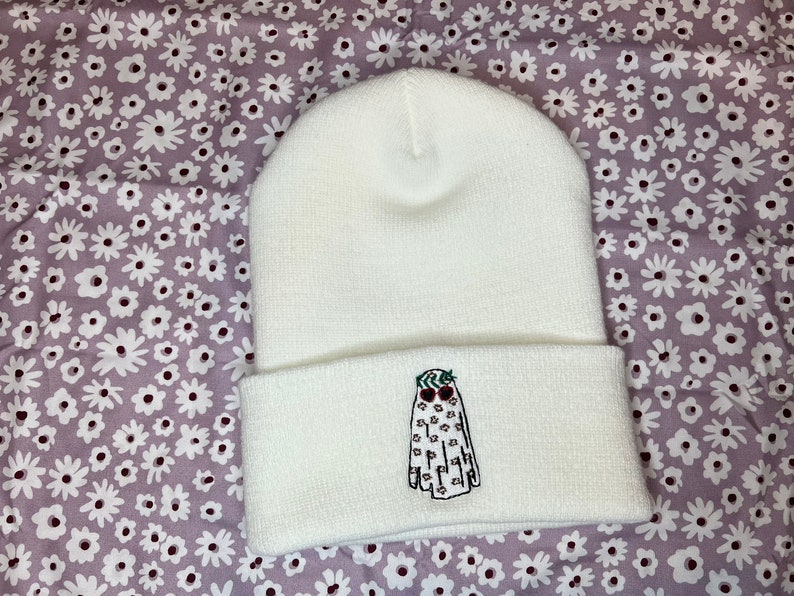 Pode incluir: Bon&eacute; branco com um fantasma bordado em branco que usa uma coroa de folhas verdes e bagas vermelhas. O fantasma tem olhos pretos e uma boca.