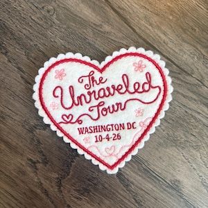 May include: A heart-shaped white felt patch with a scalloped edge and red embroidered border. It features the red text The Unraveled Tour, WASHINGTON DC, and 10-4-26, accented with small embroidered pink flowers and decorative line work.