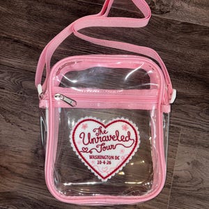 May include: A clear crossbody bag with pink trim and a matching pink adjustable shoulder strap. A white heart-shaped patch is centred on the front, featuring red text that reads The Unraveled Tour, WASHINGTON DC, and the date 10-4-26.