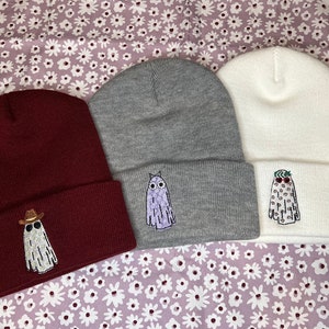 Pode incluir: Tr&ecirc;s gorros de malha em tons de vinho, cinza e branco com desenhos de fantasmas bordados. Cada fantasma tem uma express&atilde;o e um acess&oacute;rio diferente, incluindo &oacute;culos de sol, um chap&eacute;u de cowboy e uma flor.