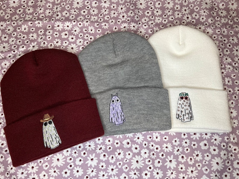 Pode incluir: Tr&ecirc;s gorros de malha em tons de vinho, cinza e branco com desenhos de fantasmas bordados. Cada fantasma tem uma express&atilde;o e um acess&oacute;rio diferente, incluindo &oacute;culos de sol, um chap&eacute;u de cowboy e uma flor.