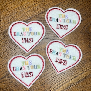 May include: Four white felt heart-shaped patches with red stitching. Each patch has the text "THE ERAS TOUR" in colorful letters and a date in black letters. The dates are 5/14/23, 5/12/23, 5/12/23, and 5/26/23.