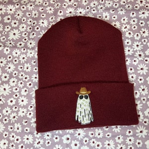 Pode incluir: Um gorro de l&atilde; cor vinho com um fantasma branco usando um chap&eacute;u de cowboy e &oacute;culos de sol bordados na frente.