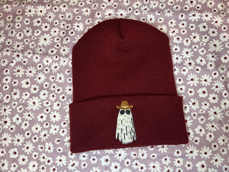 Pode incluir: Um gorro de l&atilde; cor vinho com um fantasma branco usando um chap&eacute;u de cowboy e &oacute;culos de sol bordados na frente.