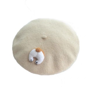 May include: A beige beret with a whimsical dog tail design. The tail is white with a brown spot and tiny black paws. The beret is a classic, round shape, perfect for adding a playful touch to any outfit.