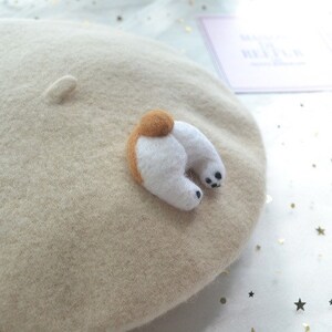 May include: A beige wool beret with a felted dog butt applique. The applique features a brown tail and white legs with black paw pads. A small, felted detail is on the beret's crown.