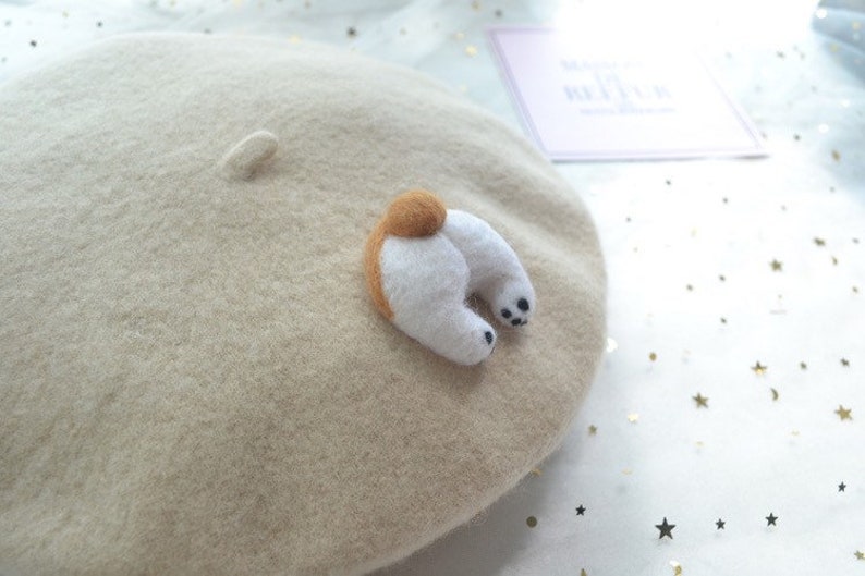 May include: A beige wool beret with a felted dog butt applique. The applique features a brown tail and white legs with black paw pads. A small, felted detail is on the beret's crown.