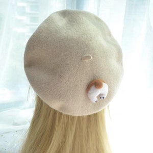 May include: A beige beret with a small, whimsical dog tail detail. The dog tail is white with a brown spot and black accents. The beret is made of a soft, felt-like material.