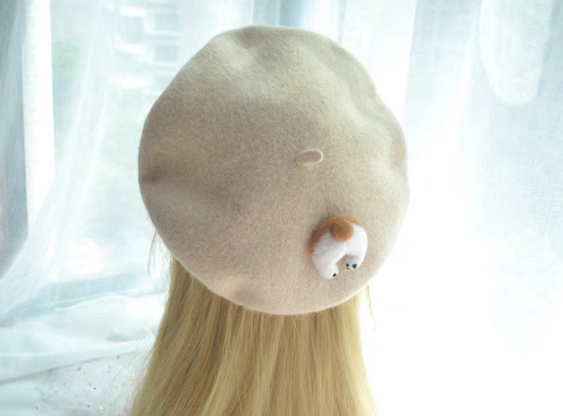 May include: A beige beret with a small, whimsical dog tail detail. The dog tail is white with a brown spot and black accents. The beret is made of a soft, felt-like material.