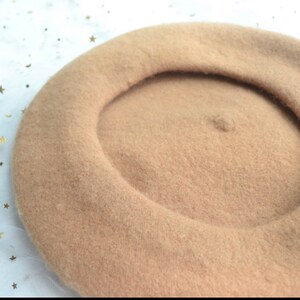 May include: A tan beret made of soft wool. The hat has a classic round shape with a defined crown. The beret is a neutral color, suitable for various outfits. Two pink heart-shaped decorations are visible in the background.
