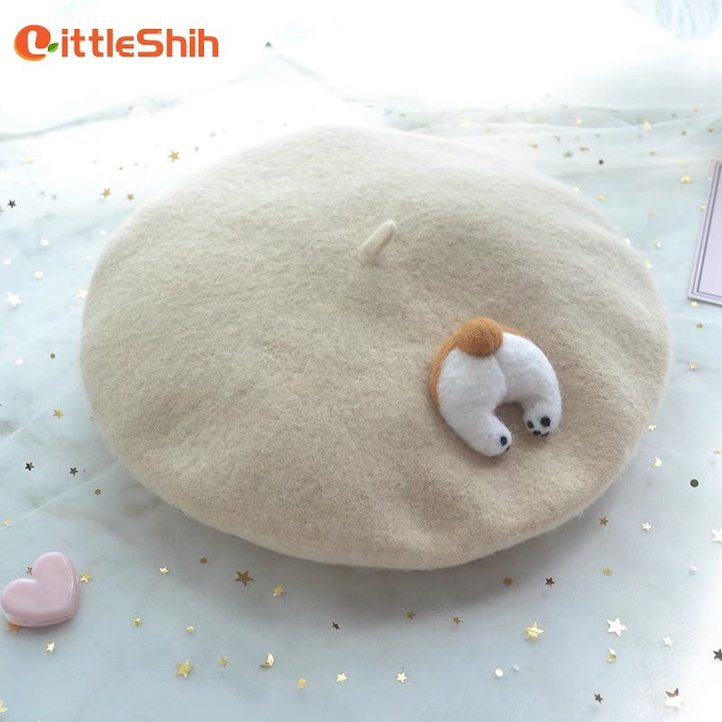 May include: A beige beret with a whimsical dog butt applique. The beret is made of a soft, felt-like material. The dog butt is white with a brown patch and black paws. The LittleShih logo is in the upper left corner.