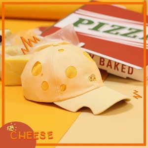 May include: A yellow baseball cap with a Swiss cheese design and a small pizza slice embroidered on the front. The cap has a curved brim and a mesh back. The text "CHEESE" is printed on a red circle in the lower left corner of the image.