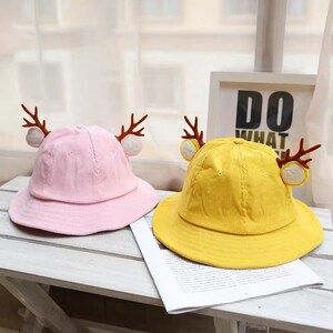 May include: Two children's bucket hats with reindeer antlers. One hat is pink and the other is yellow.